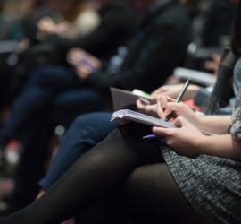A series of people sitting in a conference hall writing on notebooks